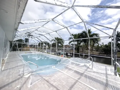Bare white aluminum pool cage frame with no screen panels installed, plastic sheeting covering the pool and paver deck
